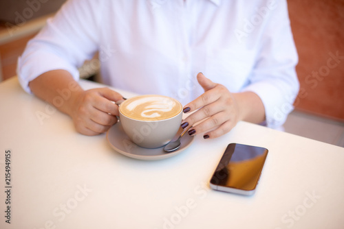 woman hands holding cup of cappuccino with latte art sitting at cafe table with mute smartphone