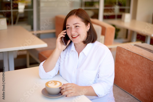 happy young woman talking on smartphone and drinking coffee sitting in outdoor cafe