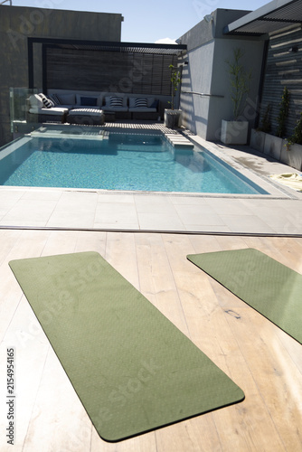 Rectangular pool is reflecting pergola shadows on clear water, olive-green mats on rooftop deck