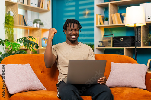 African American young man on home sofa reacts to good result on laptop with joy surprise and celebration. Black guy on home sofa smiles at screen after win in living room with excited happy emotion.