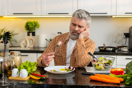 Canvas Print Middle-aged man grimacing while chewing vegetable salad in kitchen showing deep diet pressure and stress