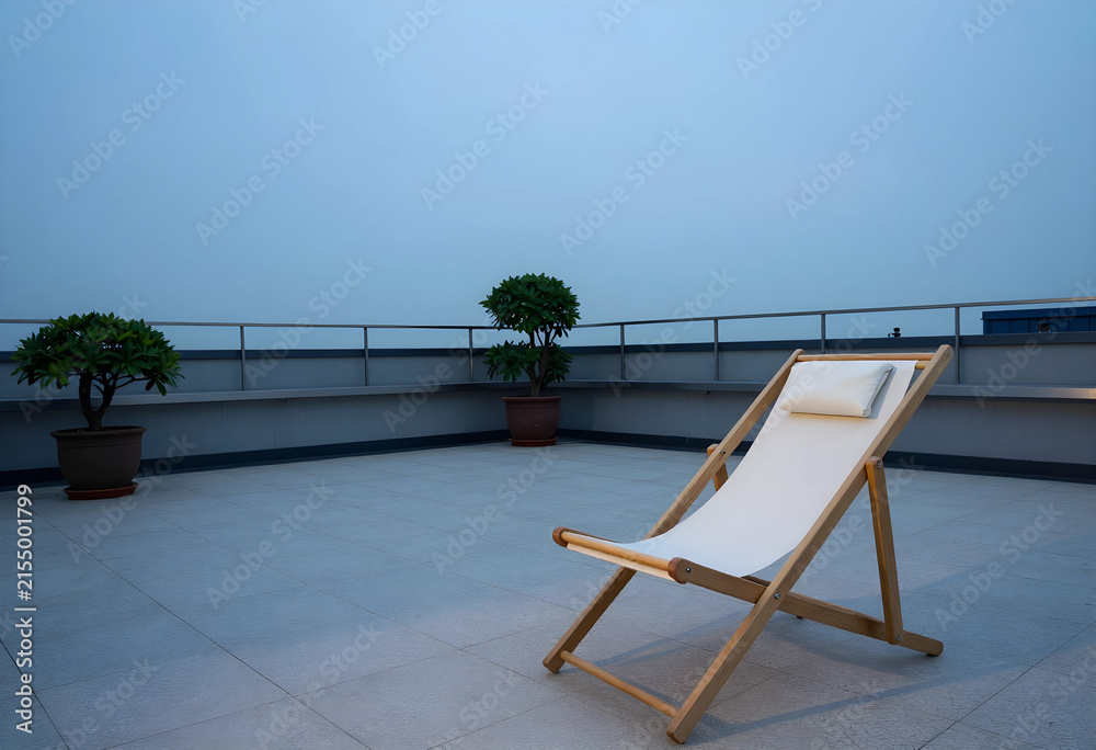Fototapeta premium Empty wooden deck chair with white fabric and pillow on a rooftop terrace with Potted plants and a railing, Potted green plants are arranged along a low wall with a metal railing