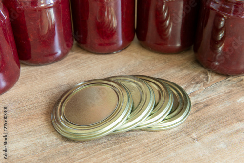 Quadro su tela Close-up of several metal canning jar lids arranged on a wooden table, with glass jars of homemade preserves softly blurred in the background