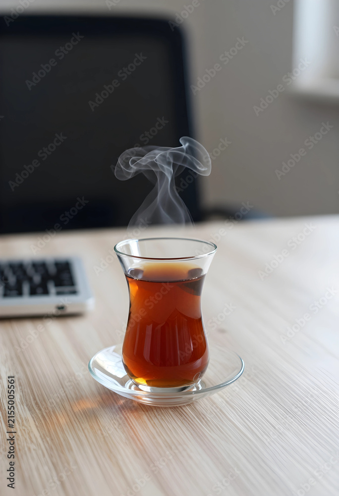 Fototapeta premium Steaming turkish tea in a traditional glass cup on a wooden desk with a laptop keyboard in the background, a laptop keyboard is visible in the blurred background, a work or study environment