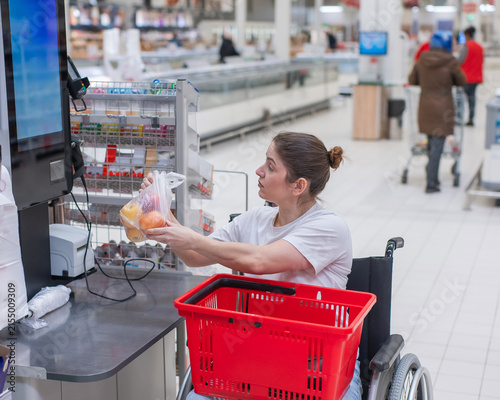 A Caucasian woman in a wheelchair pays for groceries at a self-checkout counter in a supermarket.