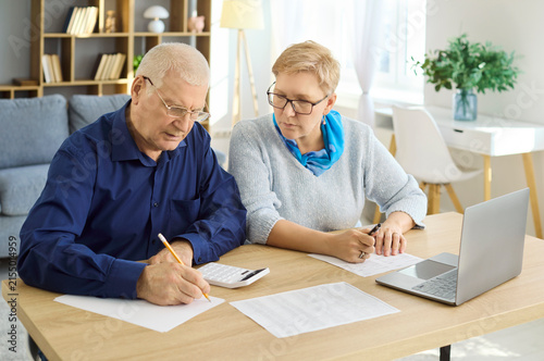 Seniors couple budgeting bills with calculator laptop. They review paperwork and verify payments for accurate finance and retirement planning. Concept of household budgeting and savings.