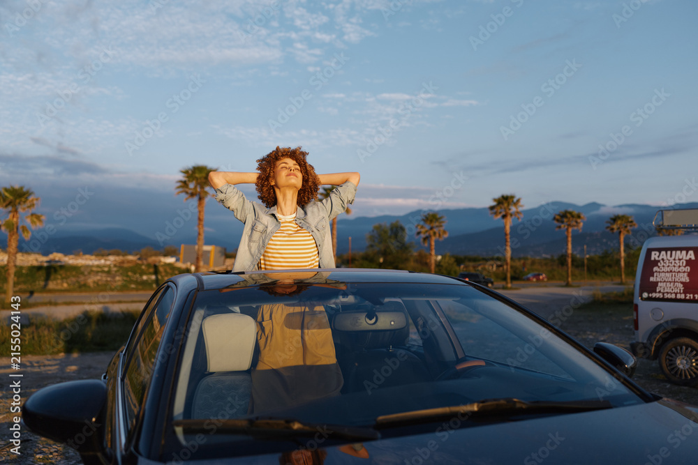 Obraz premium A woman leans on a car roof at sunset, arms behind her head, enjoying a relaxed roadtrip moment. Outdoors with palm trees and a warm sky, this lifestyle scene conveys travel and leisure.