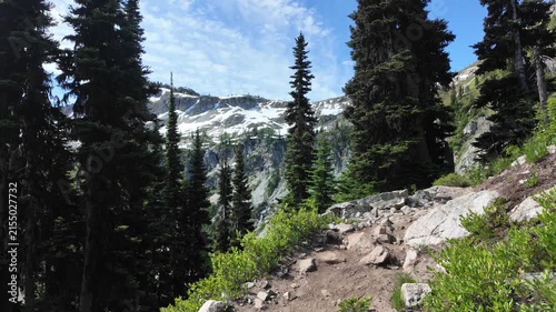 First-person point of view perspective moving forward along a dirt hiking trail surrounded by evergreen pine trees with snow-capped mountains in the background, North Cascades, Washington