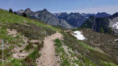 First-person point of view moving forward along a dirt mountain path with snowfield patches and dramatic mountain range in the background on a sunny day in the North Cascades, Washington