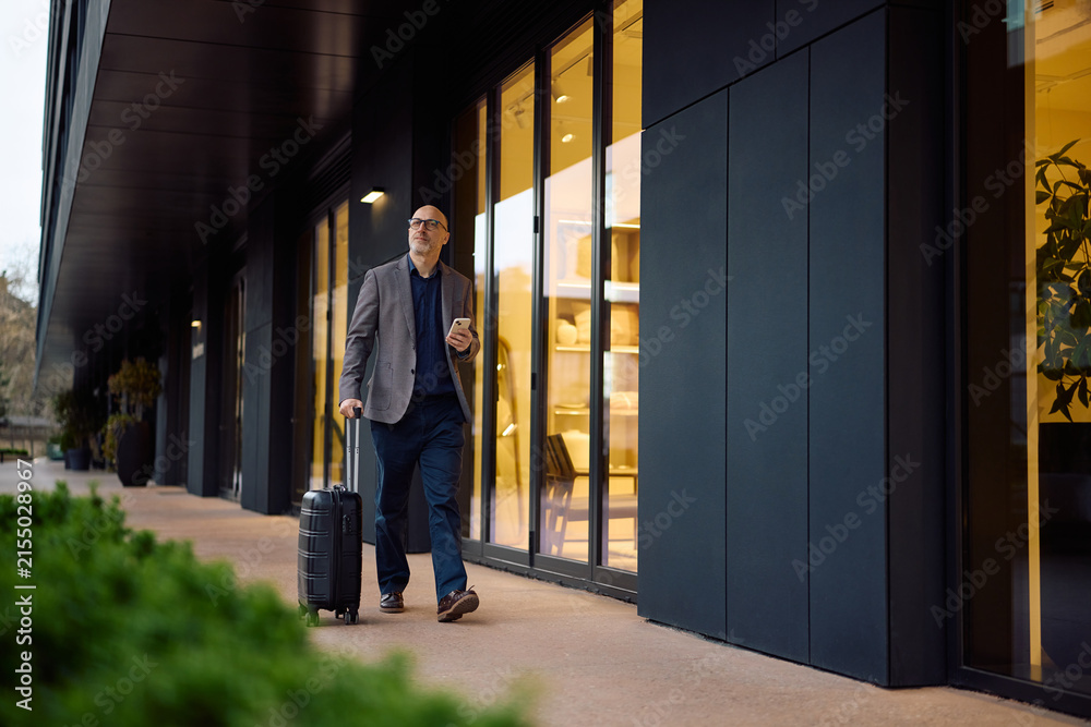 Fototapeta premium Mature businessman with smart phone and travel bag walking on street.