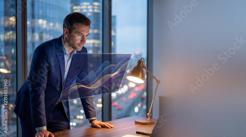 Focused businessman analyzing financial market graph on holographic screen with serious expression in office at night