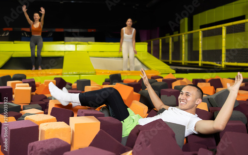 Cheerful young man enjoying soft landing in foam pit at trampoline park, with friends jumping around