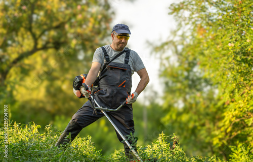 A gardener is using a weed trimmer to clear tall grass in a field. He is focused on his task, surrounded by plants and trees. The sun is shining and it is daytime