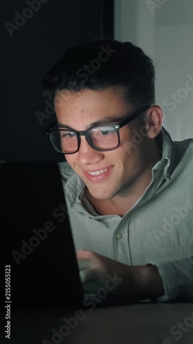 Concentrated young man wearing eyeglasses typing on a laptop computer in a dark room, with the screen light illuminating his face as he smiles, showcasing a dedicated professional working late