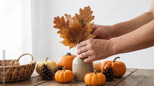 A person arranging autumn decor with pumpkins, pinecones, and dried leaves in a vase on a wooden table