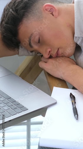 Tired young caucasian student sleeping on his desk at home after studying hard for an exam, with a laptop, a pen, and a notebook on the table. Overworked man resting due to fatigue