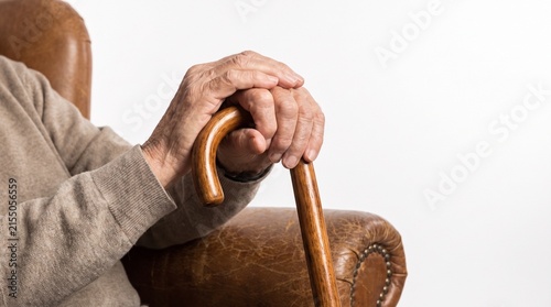 Close-up shot of an elderly person's hands resting on a wooden cane while sitting in a leather chair. The image evokes a sense of contemplation and aging gracefully.