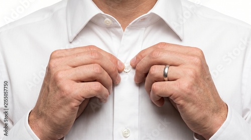 A close-up shot of a man's hands buttoning up a crisp white dress shirt, showcasing a wedding ring. The focus is on the meticulous detail of dressing for an occasion