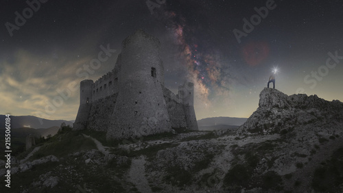 View of Rocca Calascio fortress illuminated under the Milky Way galaxy with a person standing on a nearby rock with a headlamp in Calascio, Abruzzo, Italy.