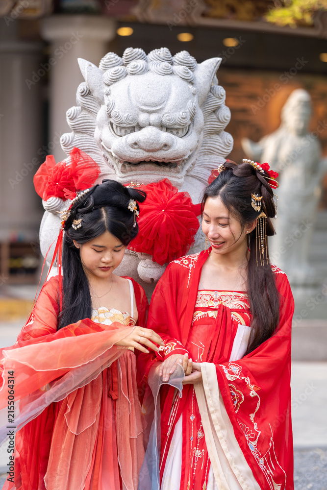 Obraz premium Wishing luck and prosperity for the Lunar New Year: two elegant young women in traditional red Chinese hanfu, hold hands, look at each other smiling, beside stone guardian lion statue with.
