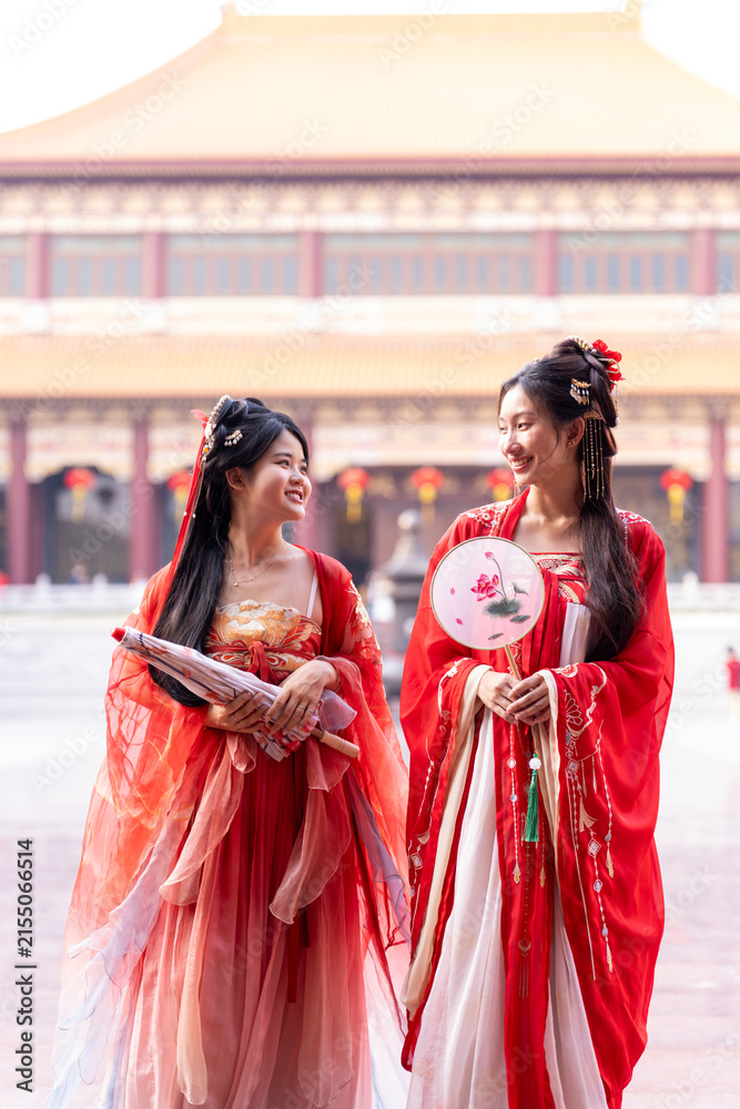 Obraz premium Wishing luck and prosperity for the Lunar New Year: two elegant young women in matching red hanfu costumes, one leaning into partner's shoulder laughing, beside temple courtyard plaza.