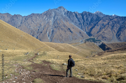 Male hiker with a backpack walks a rugged trail through a vast alpine valley in Georgia, facing massive Caucasus mountain peaks and a distant Gergeti Trinity Church under a clear blue sky