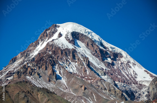 Majestic, snow capped summit of Mount Kazbek in the Caucasus range of Georgia, showcasing rugged rocky ridges and steep glacial slopes against a clear blue sky