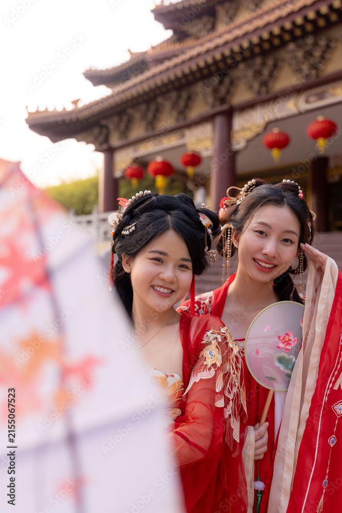 Obraz premium Portrait of two young Chinese women in red silk hanfu dresses: lowers parasol, tilts heads together smiling, framed by temple eave with red lanterns, celebrating Chinese New Year traditions.