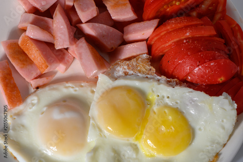 Bild auf Leinwand Brightly lit morning scene with two sunny side up eggs glistening next to juicy tomato slices and neatly sliced sausage