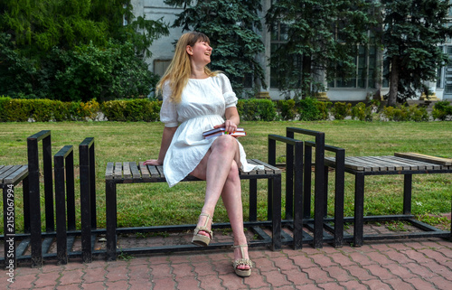 Young woman in a white dress sits on a park bench laughing and holding a stack of books, enjoying a relaxed summer day surrounded by grass, trees and an urban green space