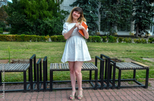 Smiling young woman in a white summer dress holds orange notebooks while standing on a park bench area, conveying student life, joy and relaxed summer vibes in a green outdoor setting