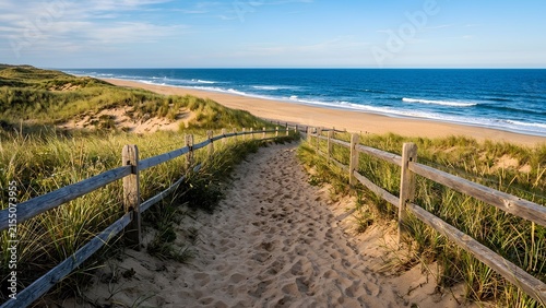 Sandy path with a wooden fence winding through grassy dunes to the ocean beach.