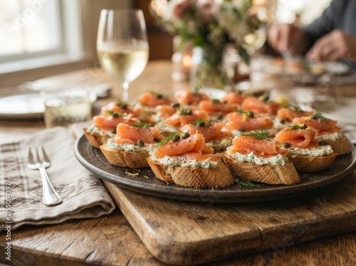A gourmet platter of smoked salmon crostini topped with cream cheese, fresh dill, and capers, served on a rustic wooden table with wine and blurred dinner setting in the background.