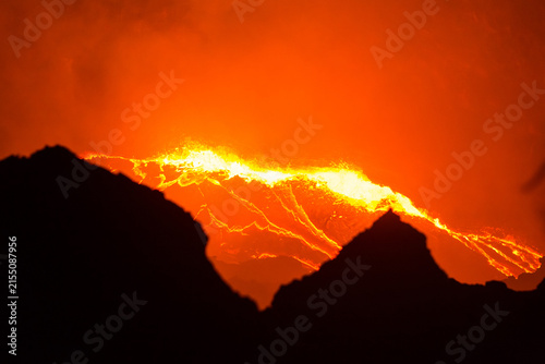 View of glowing lava flowing from the crater of Erta Ale volcano at night with dark volcanic silhouettes in the foreground, Ethiopia.