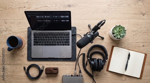Podcast Studio Setup: A neatly arranged overhead view of a home podcasting studio, featuring a laptop, microphone, headphones, audio interface, coffee, and notebook on a wooden desk.