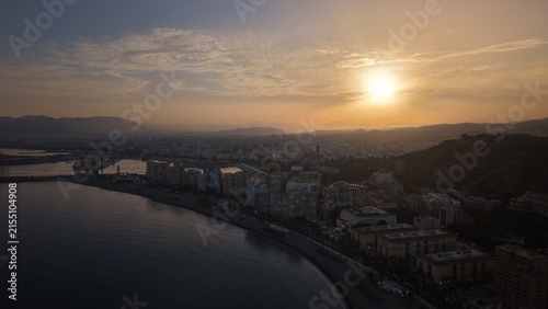 Aerial view of Malaga bullring (Plaza de Toros de la Malagueta) illuminated at sunset. Cinematic drone shot of the coastal arena, Mediterranean sea, and city skyline during golden hour.