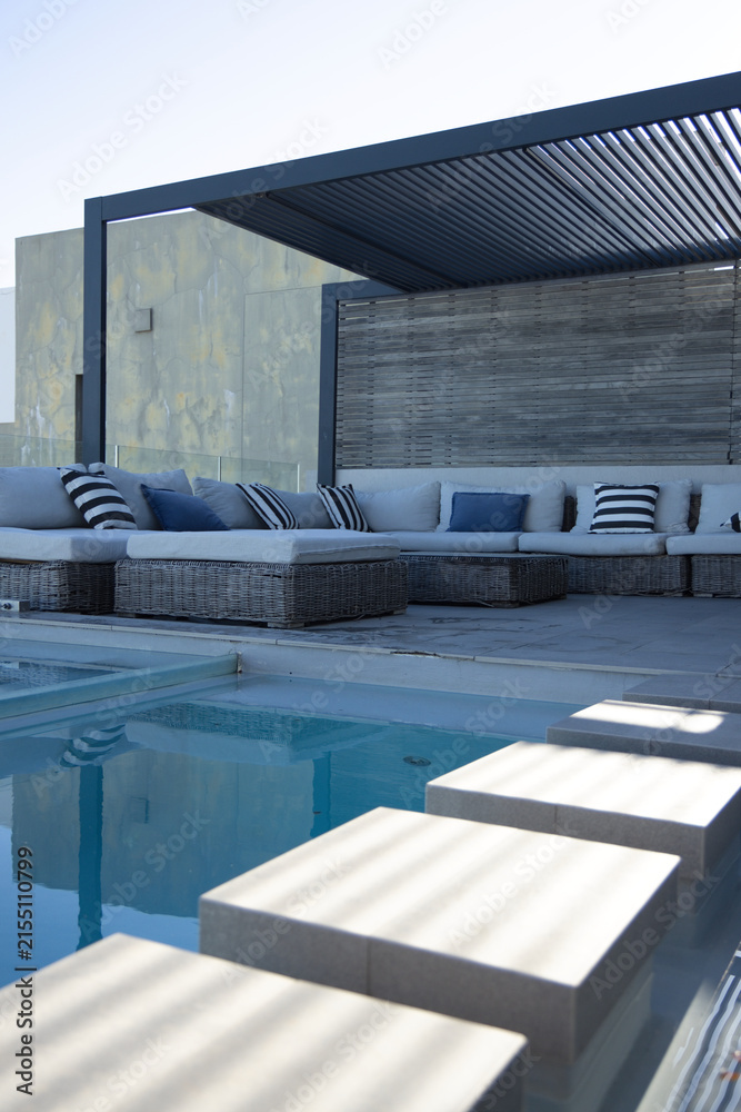 Obraz premium L-shaped rattan sectional is sitting under dark metal pergola by pool, navy pillows reflecting