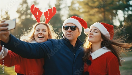 Friends taking a festive selfie outdoors in Santa hats and reindeer antlers, capturing holiday cheer and winter celebration for social media.