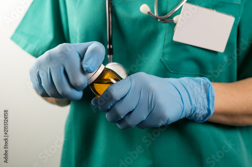 Fotografía Torso view of a medical staff member wearing green scrubs and blue nitrile gloves, carefully twisting the white cap to open an amber glass bottle filled with a yellow liquid medication