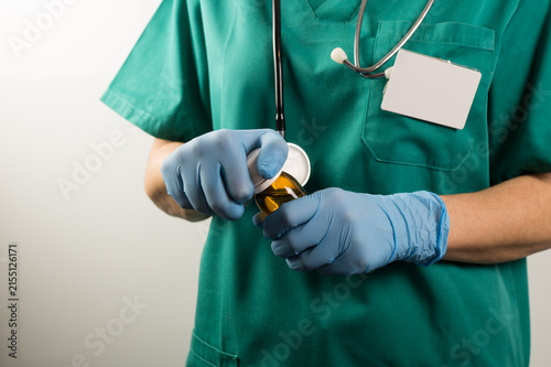 Konstfotografi Mid-shot of a medical professional wearing green scrubs, a stethoscope, and a blank ID badge, using blue gloved hands to open an amber bottle of liquid medication against a white background