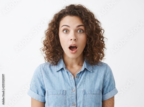 Portrait of a shocked young woman with curly brown hair looking at the camera with wide eyes and open mouth in disbelief, wearing a blue denim shirt against a plain neutral light grey background.