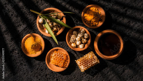 Flat lay of herbal ingredients for tropical health, clay bowl, containing golden turmeric powder, dried jasmine flowers, green pandan leaves, raw honey with natural honeycomb, and coconut oil.