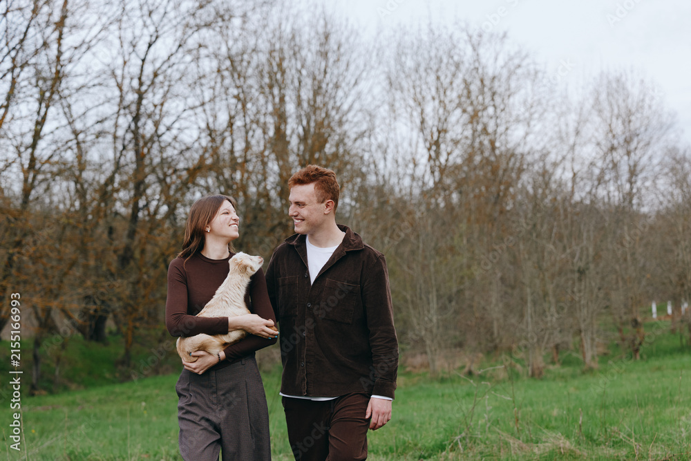 Obraz premium Casual couple outdoors with a goat kid, smiling and holding hands, in a green meadow under soft daylight, a wholesome lifestyle moment that feels natural, warm, intimate, and freely candid.