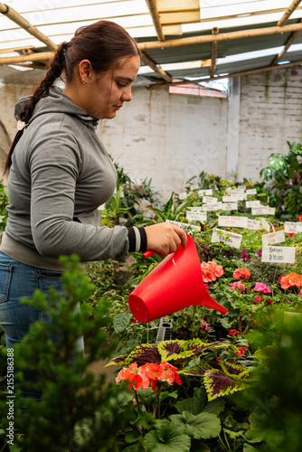 Woman nurturing various plants and flowers in a bustling greenhouse with a red watering can