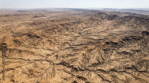 An aerial view showcases a vast, arid desert landscape. Deep, intricate ravines and ridges dominate the terrain, sculpted by centuries of erosion.