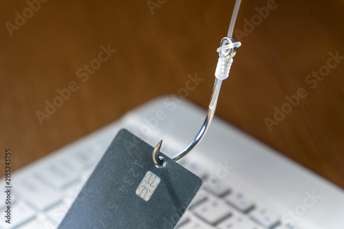 Fishing hook piercing a credit card over a blurred laptop keyboard. Online phishing scams, cybersecurity threats, digital fraud, and internet identity theft concept.
