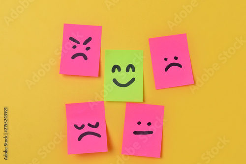 Green sticky note with a happy face surrounded by pink notes displaying negative expressions on a yellow background with copy space. Mental health concept.