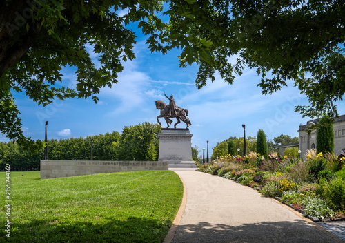 St. Louis, Missouri, United States - 07 August 2026: View of the Apotheosis of Saint Louis statue on horseback, surrounded by green grass, a gravel pathway, and flower beds under a blue sky.