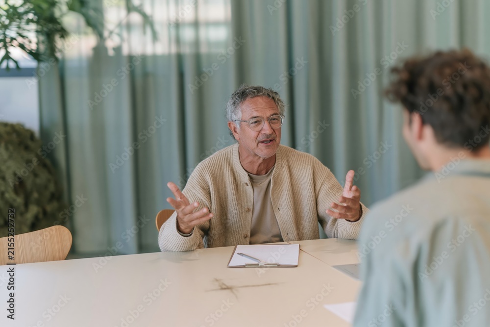 Fototapeta premium Senior man in a ribbed knit cardigan explaining a concept during a meeting with a young adult, featuring soft window light and a calm sage green background for advisory and consulting services