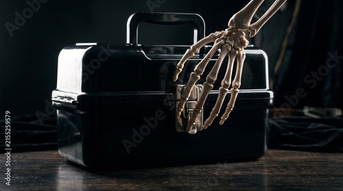 Skeleton hand reaching for black toolbox on wooden table, spooky bony fingers touching plastic tool chest latch, dark cinematic horror aesthetic for halloween mystery concept